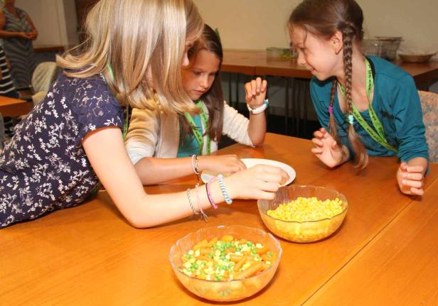 Drei Mädchen sitzen an einem Tisch mit Schüsseln mit Mais, Erbsen und Karotten.