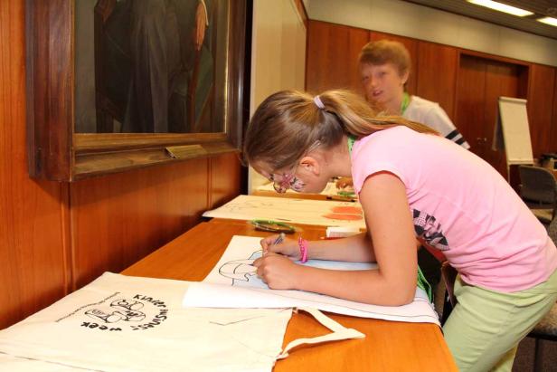 Ein Mädchen mit Gesichtsbemalung zeichnet auf ein Blatt Papier; ein Junge steht im Hintergrund.
