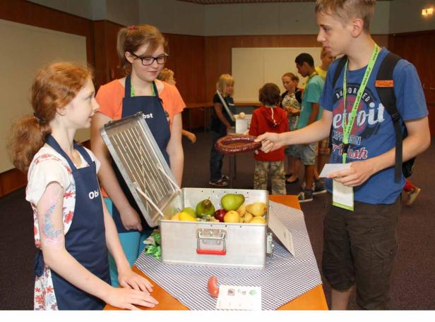 Kinder stehen an einem Tisch mit Obst und Gemüse, ein Junge hält eine Wurst in der Hand.