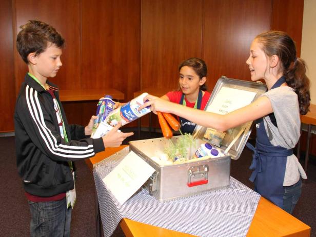 Drei Kinder befüllen eine Kühlbox mit Lebensmitteln wie Milch und Karotten.
