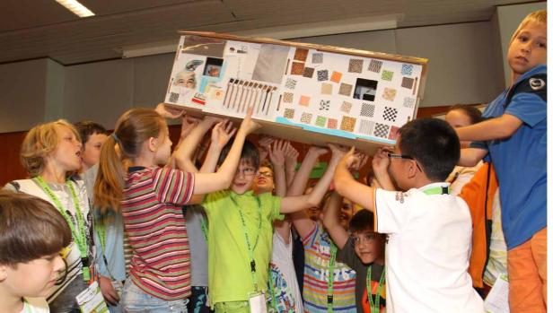 Eine Gruppe Kinder trägt gemeinsam ein großes, dekoriertes Modellhaus.