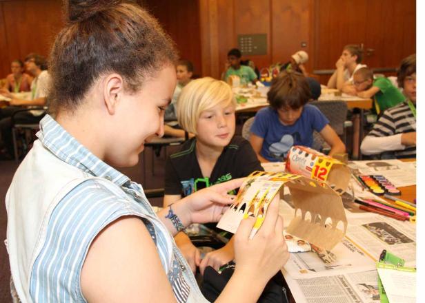 Kinder basteln in einem Raum mit Zeitungen und Malstiften an Tischen.