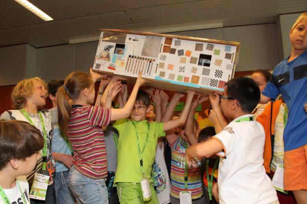 Eine Gruppe Kinder trägt gemeinsam ein großes, dekoriertes Modellhaus.