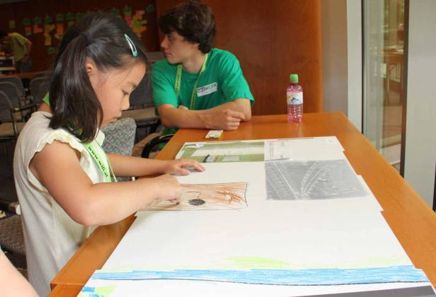 Ein junges Mädchen zeichnet an einem Tisch, während ein junger Mann zusieht.