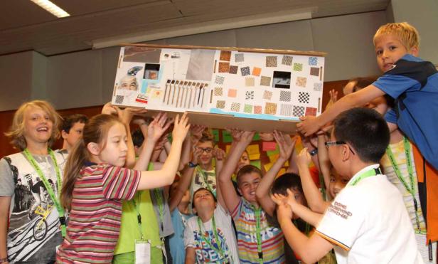 Eine Gruppe Kinder hält gemeinsam ein großes, dekoriertes Kartonhaus hoch.