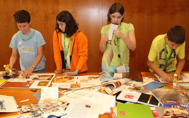 Vier Kinder basteln an einem Tisch mit Zeitschriften und Klebstoff.