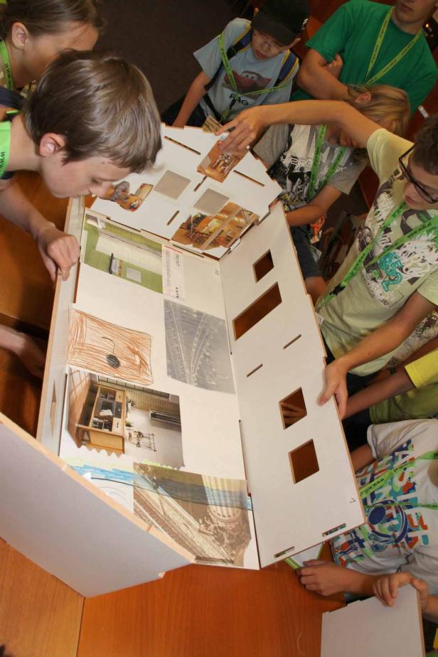 Eine Gruppe Kinder betrachtet und bearbeitet ein Modellhaus mit verschiedenen Zimmerbildern.