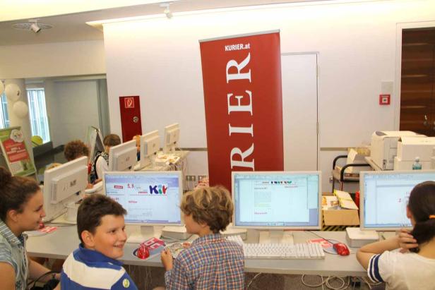 Kinder sitzen an Computern in einem Raum mit einem „Kurier.at“-Banner.