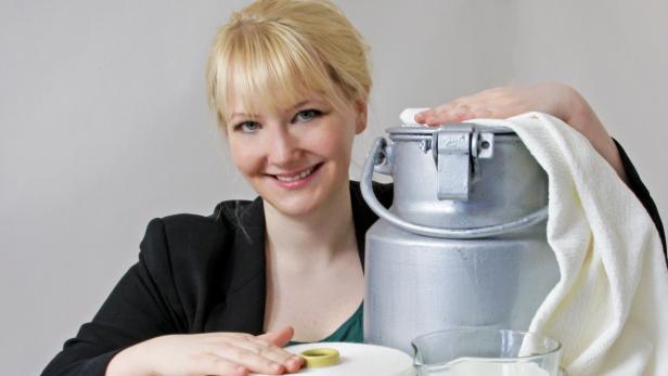 Eine lächelnde Frau posiert mit einer silbernen Milchkanne und einem Glas Milch.