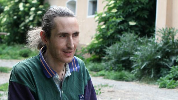 Ein Mann mit Pferdeschwanz sitzt im Freien vor Büschen.