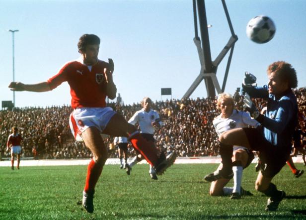 Ein Fußballspieler in Rot schießt den Ball vor einem Torwart und einer Zuschauermenge.