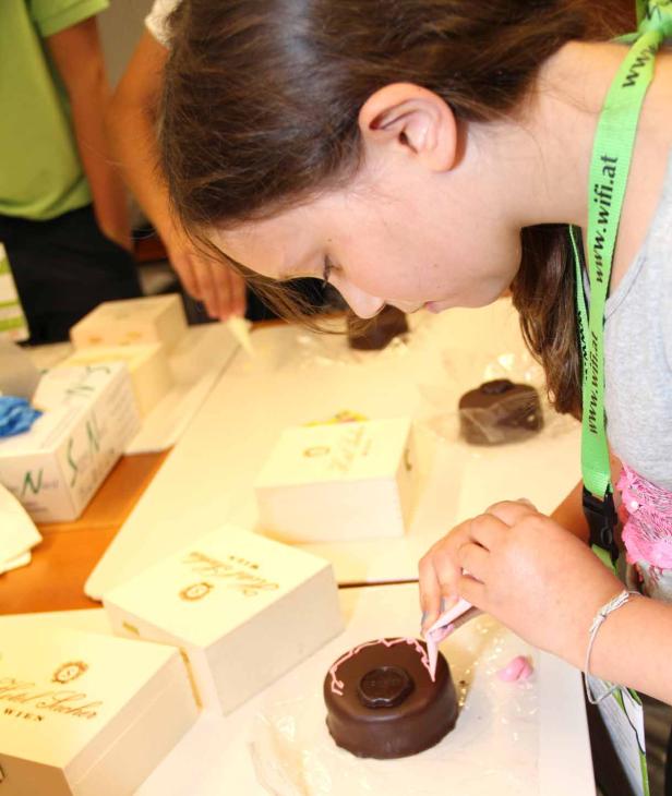 Ein Mädchen verziert eine Mini-Sachertorte mit rosa Zuckerguss.