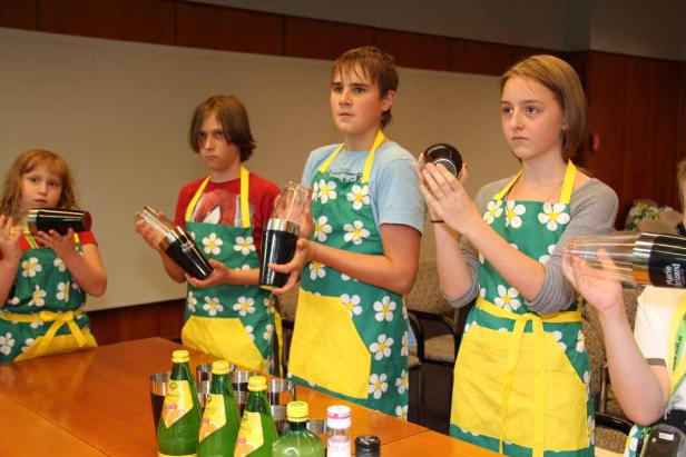 Eine Gruppe Kinder in Schürzen schüttelt Cocktails hinter einer Bar mit Zutaten.
