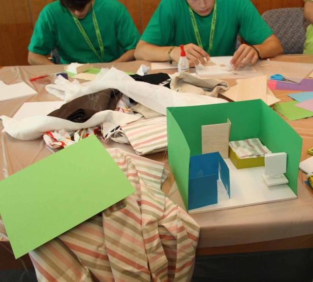 Zwei Jugendliche basteln an einem Tisch mit Stoffresten und einem Modellzimmer.