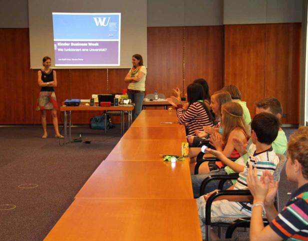 Zwei Frauen präsentieren die „Kinder Business Week“ vor einer Gruppe applaudierenden Kinder.