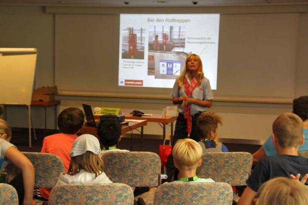 Eine Frau hält einen Vortrag über die Benutzung von Rolltreppen vor einer Gruppe von Kindern.
