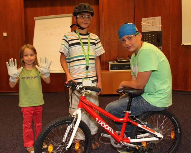 Drei Kinder posieren mit einem roten Woom-Fahrrad in einem Innenraum.