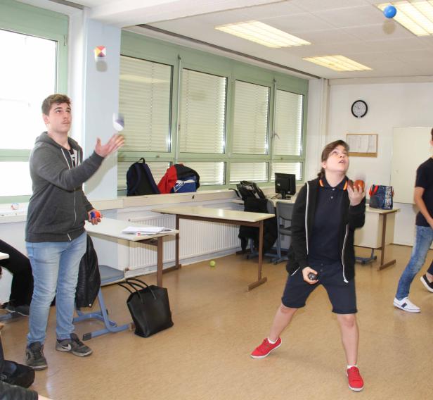 Zwei Personen jonglieren in einem Klassenzimmer mit Bällen und einem Würfel.