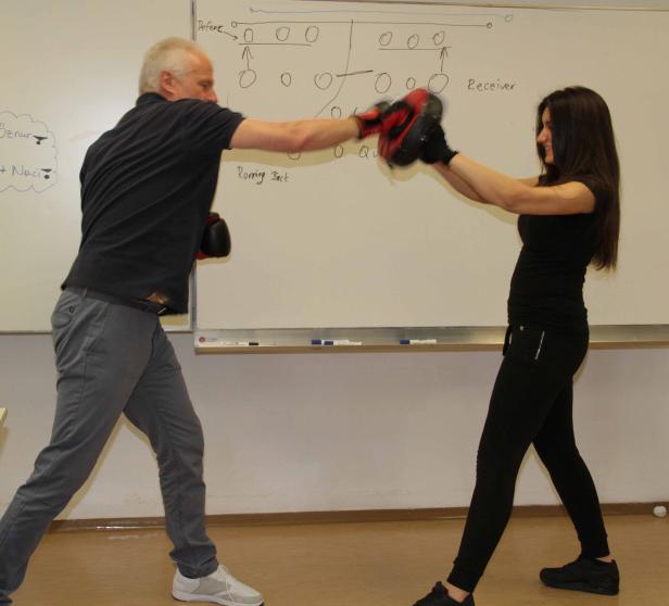 Ein Mann boxt mit einer jungen Frau vor einer Tafel mit einer Zeichnung.