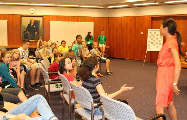 Eine Frau in einem orangefarbenen Kleid präsentiert vor einer Gruppe von Kindern in einem Konferenzraum.