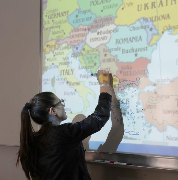 Eine Frau zeichnet mit einem Stift auf einer Europakarte die Umrisse von Griechenland.