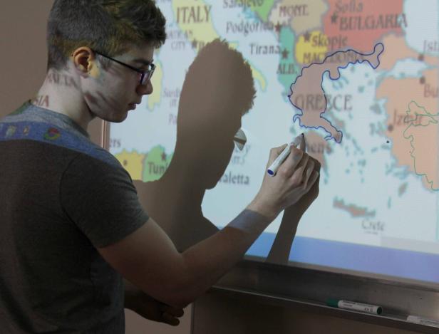 Ein junger Mann zeichnet mit einem Stift die Umrisse von Griechenland auf einer Weltkarte.