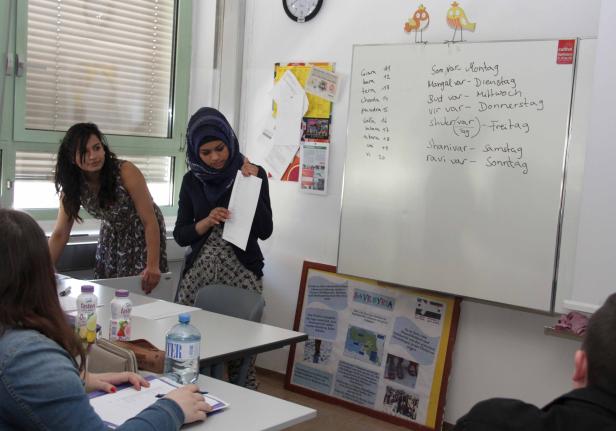 In einem Klassenzimmer stehen Schüler und eine Lehrerin vor einer Tafel mit Notizen.
