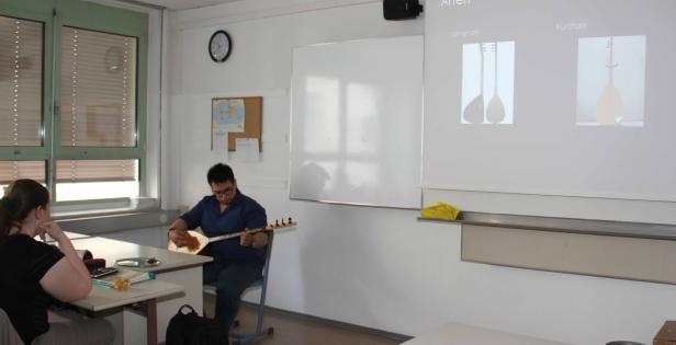 Ein Mann spielt in einem Klassenzimmer ein Saiteninstrument, während eine Frau zuhört.