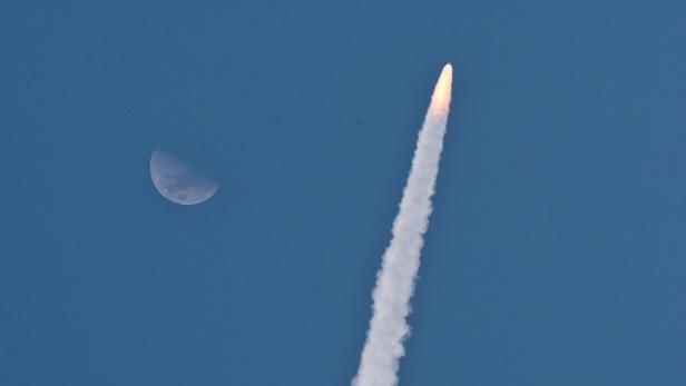 Eine Rakete steigt mit einer Rauchfahne in den blauen Himmel auf, neben dem Mond.