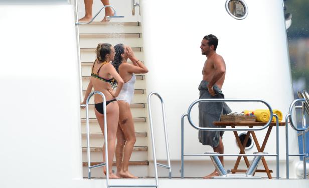 Zwei Frauen duschen auf dem Deck einer Yacht, während ein Mann mit einem Handtuch um die Hüften daneben steht.