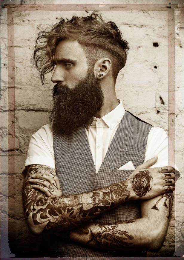 Ein Mann mit Bart und Tätowierungen steht vor einer Ziegelwand.