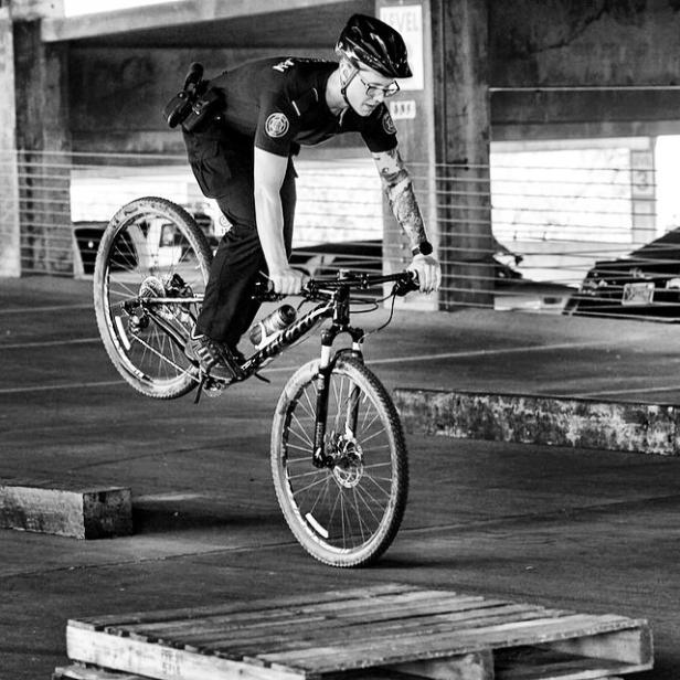 Ein Polizist fährt auf einem Mountainbike auf einem Hinterrad über eine Palette.