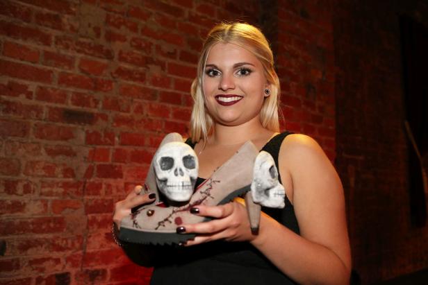 Eine Frau hält ein Paar Schuhe mit Schädeln vor einer Ziegelwand.