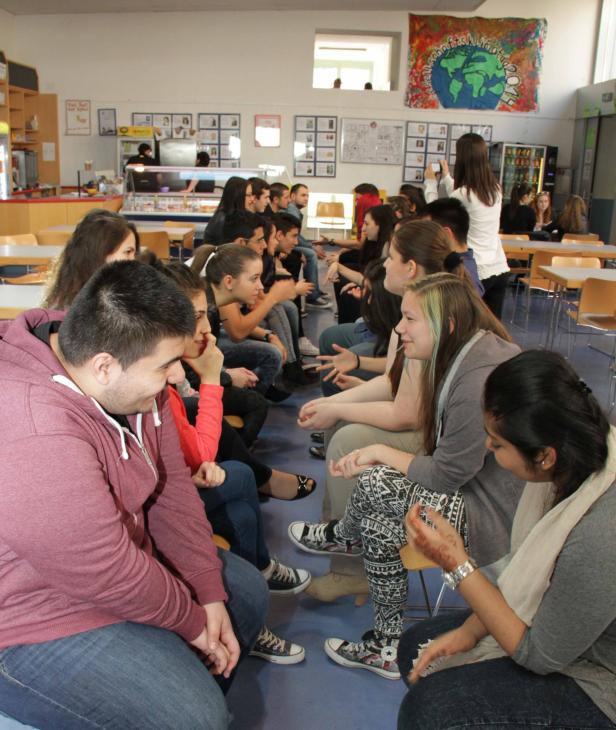Schüler sitzen in zwei Reihen einander gegenüber und diskutieren in einer Schulkantine.