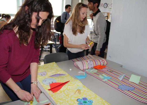 Zwei Frauen basteln an einem Tisch mit Papier und Klebstoff, während andere im Hintergrund stehen.