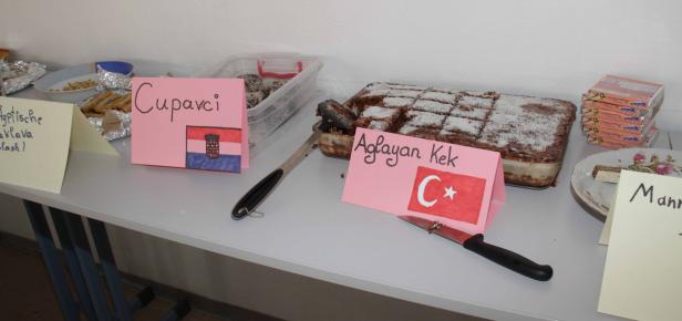 Auf einem Tisch stehen verschiedene Speisen, darunter „Cupavci“, „Aglayan Kek“ und „Manner Neapolitaner“.