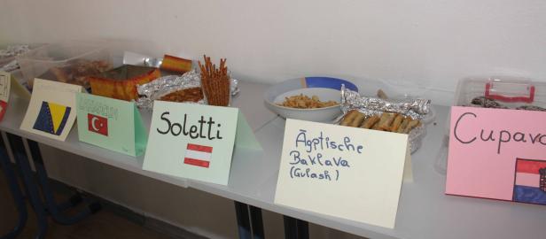Ein Tisch mit verschiedenen Speisen und handgeschriebenen Schildern, die Cevapcici, Lahmacun, Soletti, Ägyptische Baklava und Cupava bezeichnen.