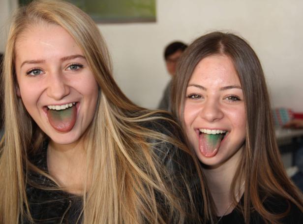 Zwei junge Frauen zeigen lachend ihre grün gefärbten Zungen.