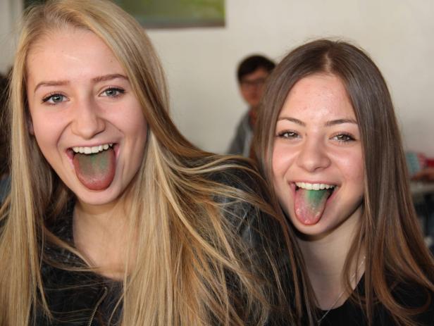Zwei junge Frauen zeigen lachend ihre grün gefärbten Zungen.