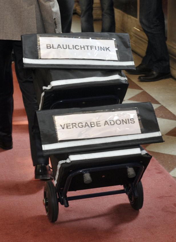 Mangelware: Akten. Ein Gepäckwagen mit der Aufschrift „Blaulichtfunk“ und „Vergabe Adonis“ wird gezogen.