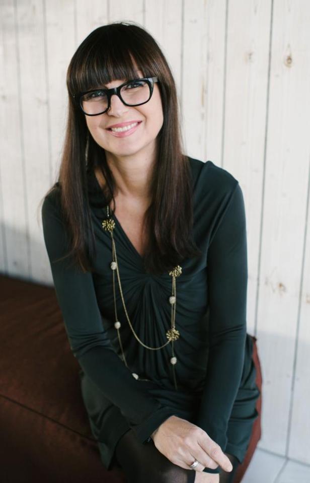 Eine lächelnde Frau mit Brille und dunklem Kleid sitzt vor einer Holzwand.