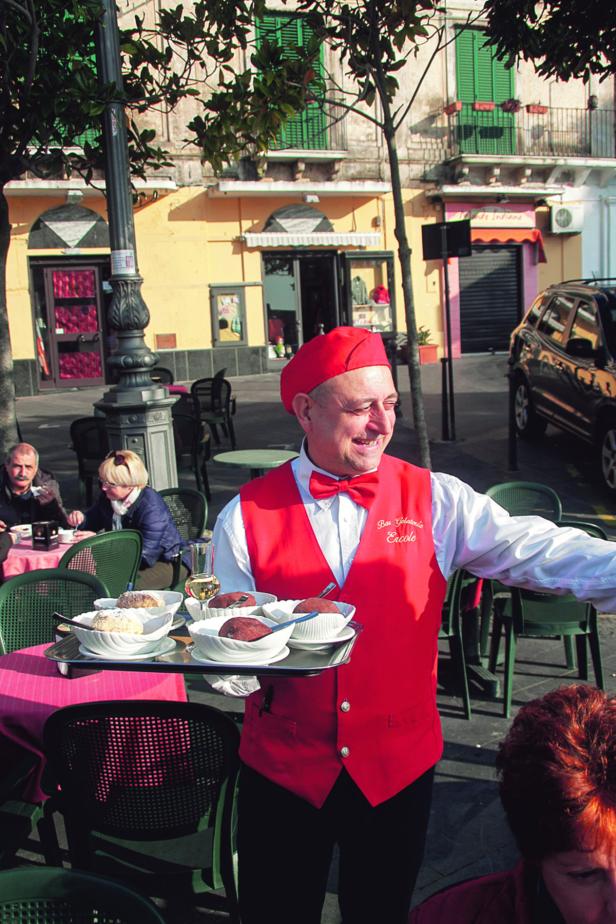 Ein Kellner in roter Weste und Barett serviert Desserts in einem Straßencafé.