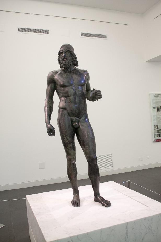 Eine Bronzestatue eines bärtigen, nackten Mannes auf einem Marmorsockel.