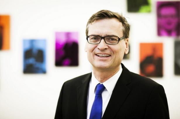 Ein lächelnder Mann mit Brille, Anzug und Krawatte vor farbigen Porträtgemälden.