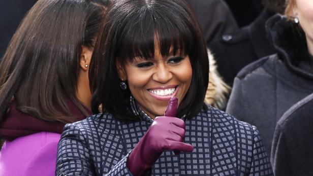 Michelle Obama lächelt und zeigt mit einem lilafarbenen Handschuh nach oben.