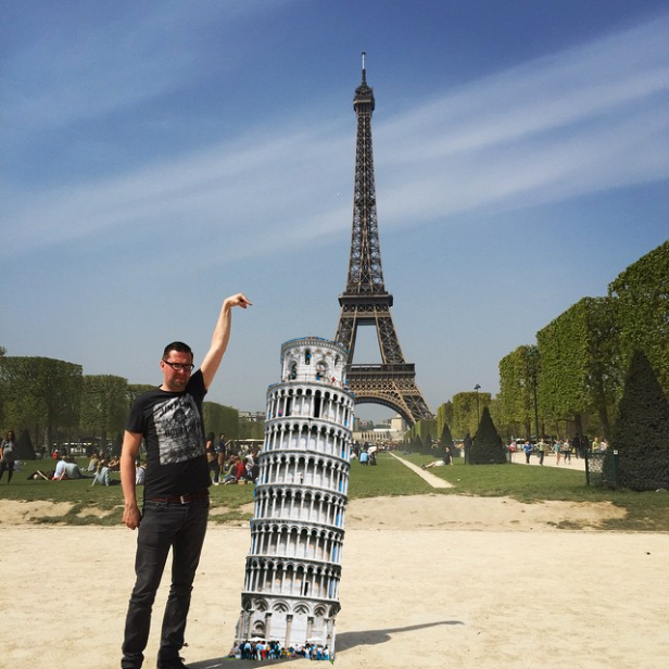 Ein Mann posiert vor dem Eiffelturm und einem Modell des Schiefen Turms von Pisa.