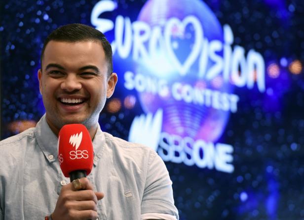 Ein lächelnder Mann hält ein rotes SBS-Mikrofon vor einem Eurovision Song Contest-Hintergrund.
