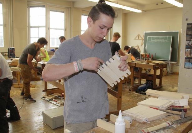 Ein junger Mann prüft in einer Werkstatt ein Holzteil mit Zinkenverbindung.