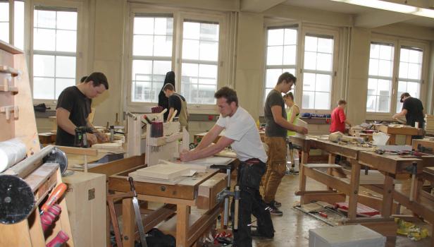 Mehrere Personen arbeiten in einer hellen Werkstatt an Holztischen.
