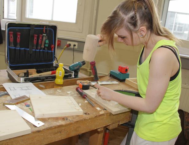 Eine junge Frau arbeitet mit Stecheisen und Hammer an einem Werkstück aus Holz.